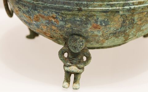 Close-up of a weathered bronze vessel and its small human-shaped decorative foot, showing green and brown surface patina.