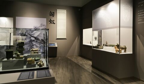 Dark exhibition gallery with illuminated cases containing Han-dynasty architectural models, vessels and figurines, accompanied by bilingual interpretation.
