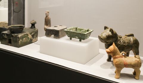 Display case containing glazed architectural and household models, a standing figure, and ceramic pig and dog figurines from the Han dynasty.