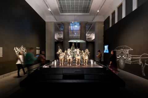 Dark exhibition gallery centered on a four-horse ceremonial chariot, with line drawings of ancient Chinese chariots on the walls.