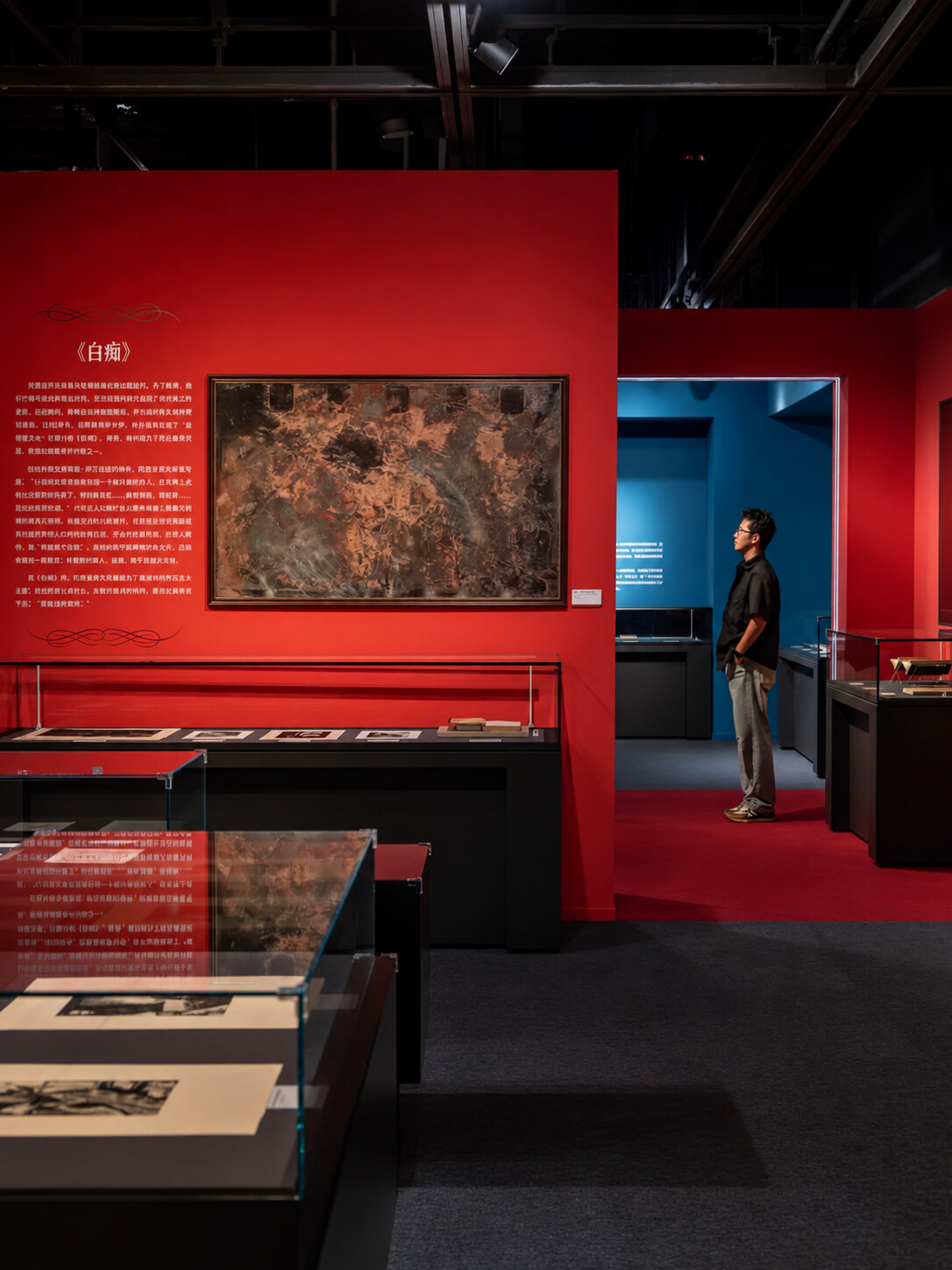 Installation view of the exhibition “Dostoevsky’s World: To Stop Reading Is to Stop Thinking,” showing a red gallery section on The Idiot with a framed artwork, display cases of printed materials, and a visitor standing near a blue adjoining room.
