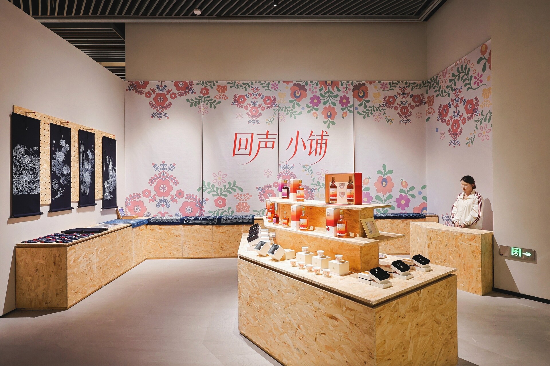 “多瑙河畔的生活之歌：匈牙利非物质文化遗产展”杭州展厅现场，可见木质展台、民俗花卉纹样墙面、黑色织物展品、瓶装展品和小型陈列物，右侧有一位坐着的观众或工作人员。