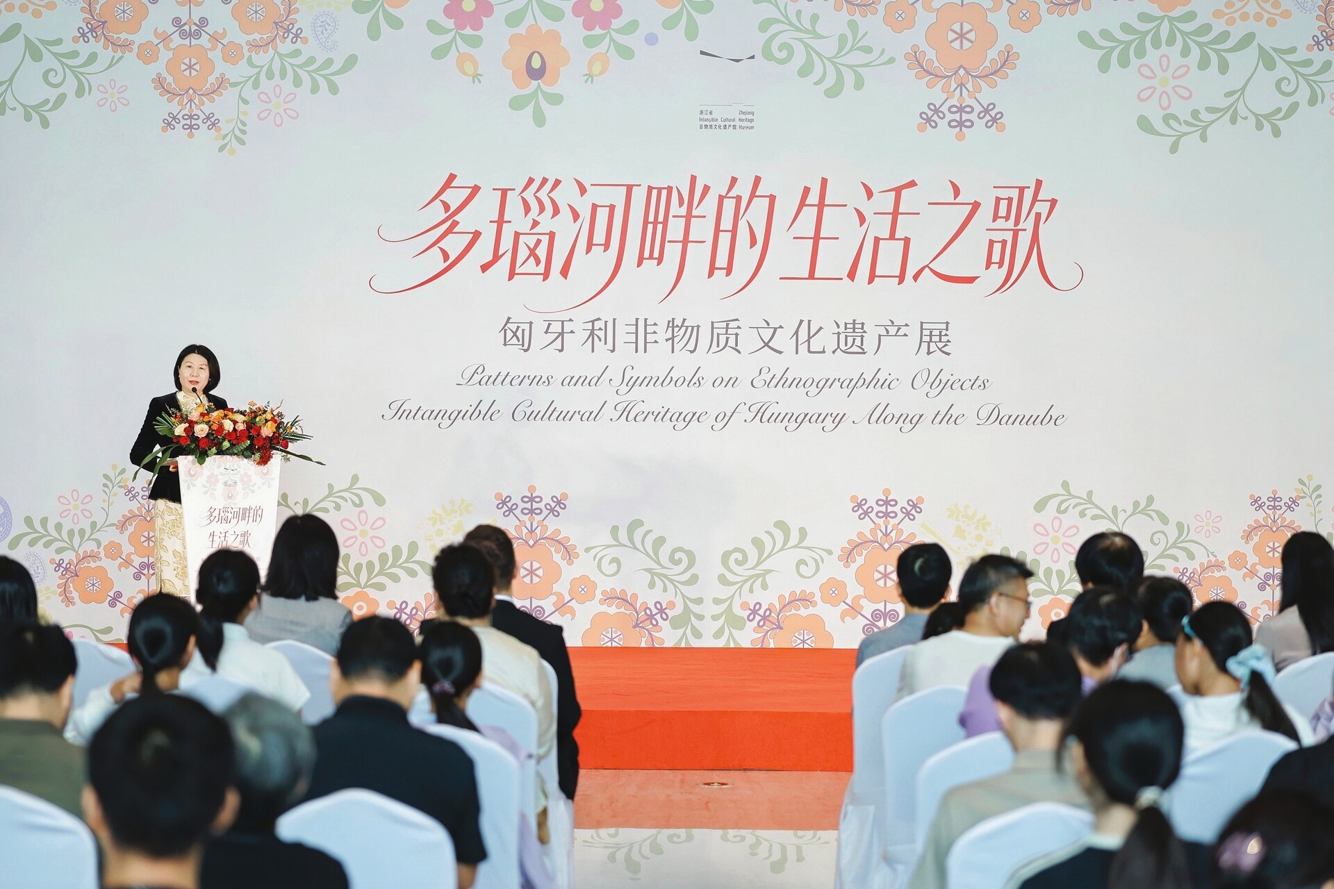 郭艺主任在装饰有鲜花的讲台前向观众致辞，背景板为浙江省非物质文化遗产馆“多瑙河畔的生活之歌：匈牙利非物质文化遗产展”的花卉图案展览主视觉。